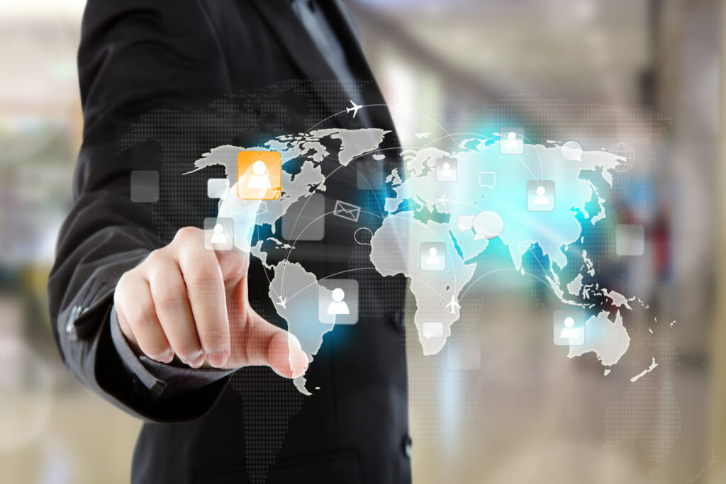 pessoa em traje social tocando uma tela virtual transparente com um mapa-múndi sobreposto. Ícones de perfil (representando pessoas ou conexões) estão distribuídos por diversos continentes, conectados por linhas, aviões e ícones digitais (como e-mails e redes). Um dos ícones está em destaque, sugerindo a seleção de um contato, oportunidade ou vagas internacionais.