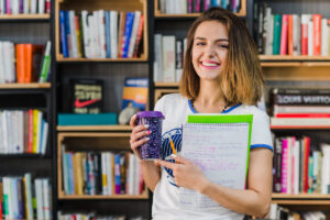 Mulher sorridente segurando caderno com anotações e copo térmico em biblioteca, simbolizando preparação para teste de proficiência em inglês.